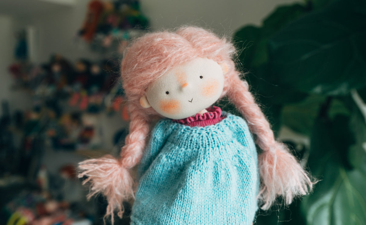 A handmade soft doll with pink mohair hair, a knitted light blue hand knit dress, and a white face with rosy cheeks, photographed in a cozy indoor setting with a blurred background of plants and other toys.